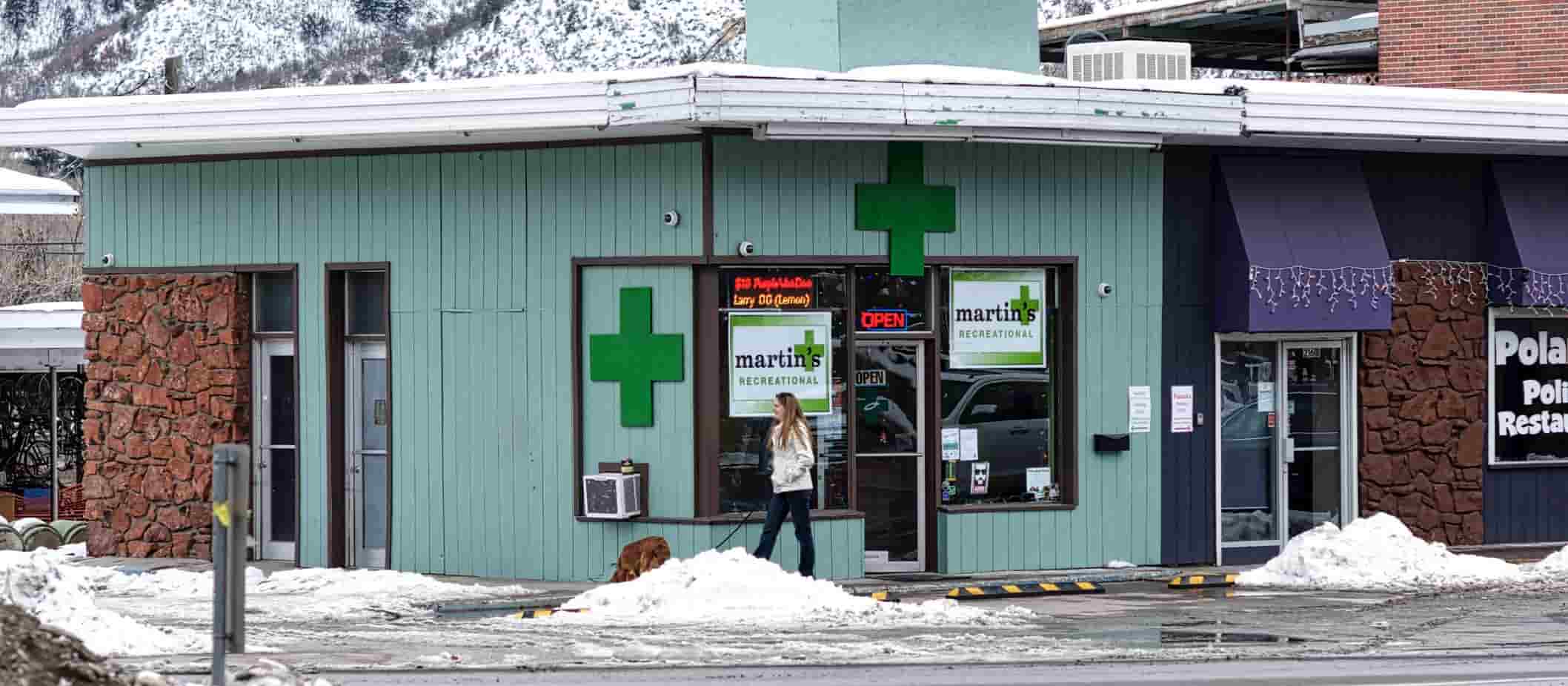 Glenwood Springs, Colorado, USA - January 6, 2016: A woman and her dog stroll past the tell-tale green crosses on a storefront in downtown Glenwood Springs that identifies it as a legal drug shop.